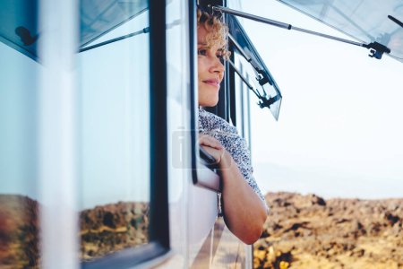 Photo for Nice adult woman smile and enjoy freedom at the natural campsite admiring outdoors outside the window of her camper car. Concept of summer tourist travel vacation and free female independence people - Royalty Free Image