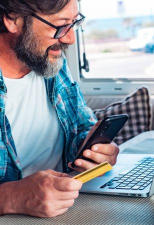 Photo for Digital nomad and remote worker freedom lifestyle concept. Adult man buying online using laptop and mobile phone connection inside a camper van in travel. Modern people and alternative office job - Royalty Free Image