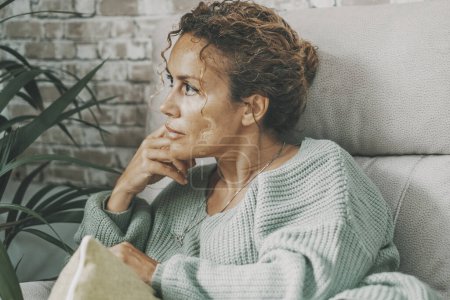 Foto de Thoughtful and worried lady at home sitting on the sofa looking serious and thinking a lot. Female portrait indoor. Lady sits on the couch touching his face with problems in mind. Finding ideas concept - Imagen libre de derechos