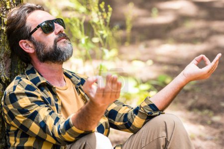 Photo for Adult  bearded man meditate and have relaxed outdoor leisure activity in the forest woods sitting against a tree trunk and enjoying nature feeling and love around. Male people enjoy life - Royalty Free Image