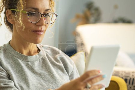 Photo for Close up portrait of modern woman female adult people reading book from electronic reader device tablet. Lady wearing eyeglasses and smile enjoying relax time at home sitting on the sofa - Royalty Free Image