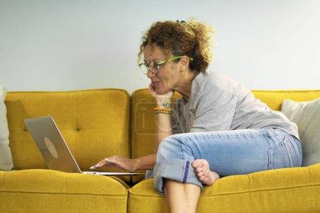 Téléchargez les photos : Femme intelligente de travail moderne en utilisant un ordinateur portable à la maison confortablement assis sur le canapé. Le travailleur à distance utilise la technologie de connexion Internet sur le canapé du hall d'hôtel - en image libre de droit