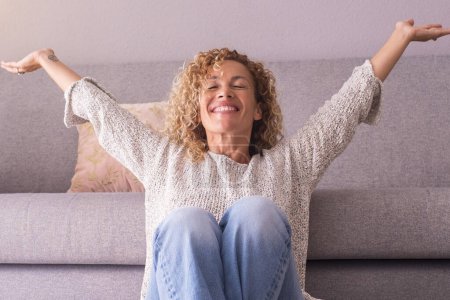 Photo for Happy and satisfied woman at home sitting on the floor, opening arms and smiling. Concept of joyful lifestyle, female people. Indoor leisure activity, Success and enjoying lifestyle. Indoor apartment - Royalty Free Image