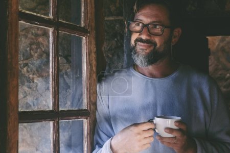 Photo for Mature man at home smile and enjoy indoor relax drinking tea or coffee and looking outside the window. People in winter chalet vacation room. Handsome male smile and think relaxing indoor - Royalty Free Image