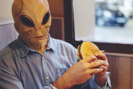 Foto de Alien con ropa humana comiendo hamburguesa dentro de una comida rápida sentado a la mesa solo. Concepto de invasión extraterrestre e inmigratoria. Nutrición de comida chatarra poco saludable - Imagen libre de derechos