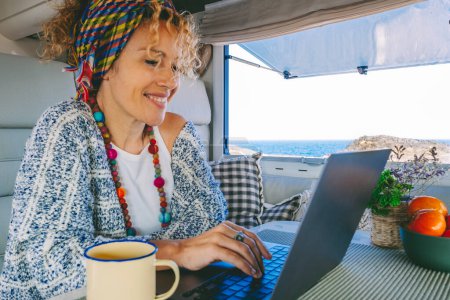 Foto de Mujer moderna de trabajo en el ordenador portátil dentro de una caravana van rv con vista al mar hermoso océano exterior. Concepto de trabajo de libertad en línea y gente trabajadora inteligente. Nómada digital usando conexión - Imagen libre de derechos