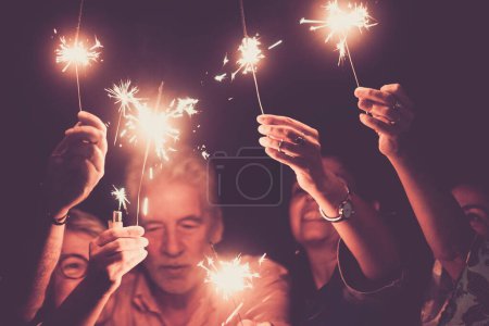 Téléchargez les photos : Groupe de personnes d'âges mixtes célèbrent ensemble le Nouvel An ou fête fête. Famille amusez-vous avec des étincelles de lumière la nuit. Amis profiter de Noël en arrière-plan noir. Célébration Noël - en image libre de droit
