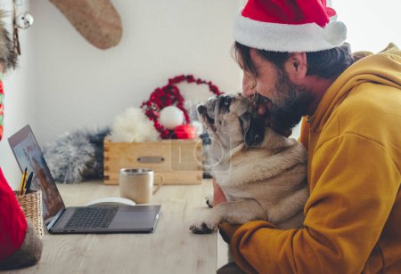 Téléchargez les photos : Noël fête à la maison avec un seul homme en vidéoconférence sur ordinateur portable étreignant son adorable chien animal de compagnie. Concept de personnes appréciant décembre à la maison avec des décorations de Noël et meilleur ami - en image libre de droit