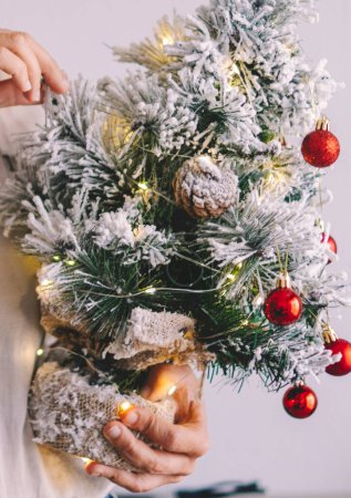 Téléchargez les photos : Femme tenant un petit arbre de Noël plein de décorations. Vacances nouvel an Noël jour de la saison. Les femmes méconnaissables profitent de la veille de Noël seules dans les loisirs à la maison intérieure. Fête - en image libre de droit
