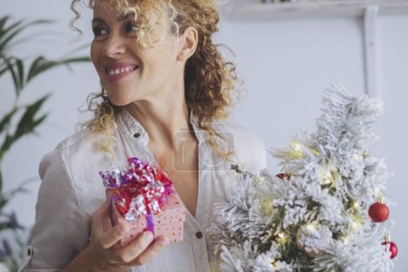 Téléchargez les photos : Joyeux sourire femme et tenir un petit arbre de Noël et cadeau boîte cadeau cadeau. Les femmes profitent de Noël saison de la veille à la maison dans les loisirs d'intérieur seul. Personne réelle en décembre jour de style de vie - en image libre de droit