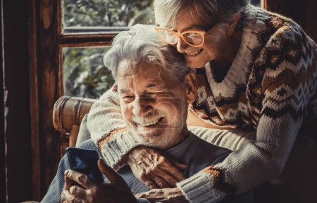 Téléchargez les photos : Heureux vieux couple utilisent téléphone portable à la maison ensemble pour appeler les parents vidéo. Saison d'hiver loisirs d'intérieur pour les retraités adultes. Femme câlin homme mûr sur la chaise. Amour et technologie - en image libre de droit