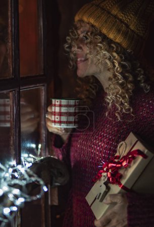 Foto de Mujer sola en casa bebiendo té en la temporada navideña celebrando regalo y mirando fuera de la ventana por la noche. Gente feliz y celebración de diciembre. Nochevieja y ambiente interior de la casa - Imagen libre de derechos