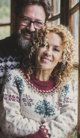 Téléchargez les photos : Portrait de couple mature souriant et debout pour une image, Homme et femme portant un pull chaud avec des décorations de Noël et de la couleur. Les amoureux dans un chalet en bois ou à la maison dans les loisirs - en image libre de droit