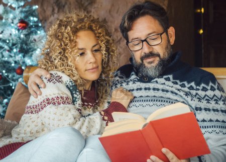 Photo for Serene  mature couple at home enjoying relax and indoor leisure activity in december holiday. Christmas tree in background. Man reading a book with a woman. Married together relationship love - Royalty Free Image