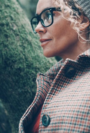 Téléchargez les photos : Portrait latéral de belle femme avec des lunettes portant une veste chaude et un chapeau en tricot. Arbres verts fond. Les femmes dans le parc naturel dans les loisirs de plein air. Une femme dans la nature. Environnement - en image libre de droit