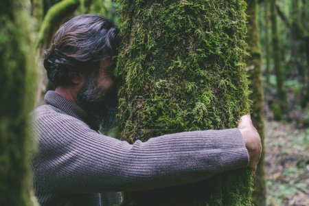 Téléchargez les photos : Vue latérale d'un homme embrassant un arbre avec du musc vert. Concept de personnes amoureux de la nature et de l'environnement. Le changement climatique et le réchauffement climatique cessent. La fête du jour de la Terre. Homme liant le tronc dans les bois forestiers - en image libre de droit