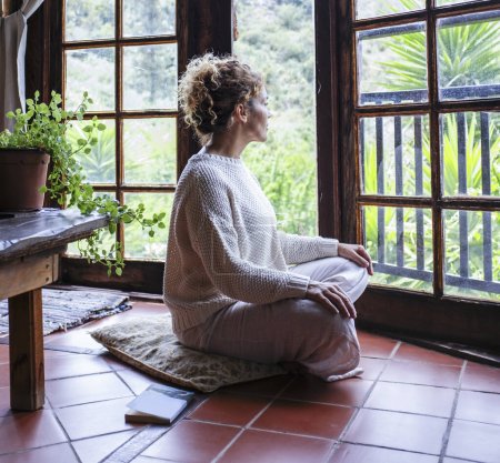 Photo for Beautiful calm caucasian woman sitting on a comfortable pillow placed on floor at home and feeling grateful and grateful. Happy woman shows gratitude, love and care, looking out the window. concept of faith and relaxation. - Royalty Free Image