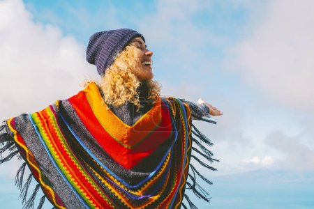 Photo for Blonde woman with hat and colorful poncho smiles and relaxes with open arms in the blue winter sky. People's freedom style. - Royalty Free Image