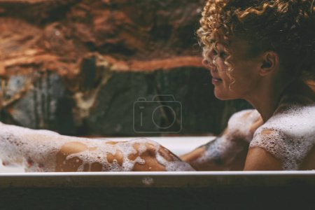 Téléchargez les photos : Vue latérale d'une femme détendue prenant un bain à la maison, allongée dans une belle baignoire, frottant sa peau avec une éponge et souriant, espace de copie. Accueil spa, hygiène, concept de relaxation - en image libre de droit