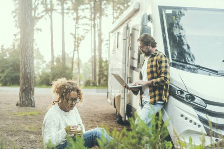 Photo for People living in rv vehicle motorhome camper van concept lifestyle. One woman having relax with plants and man working digital nomad on laptop outside. Scenic forest woods in background. Traveler life - Royalty Free Image
