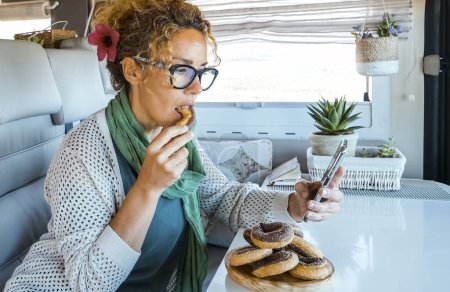 Photo for Woman eating donuts inside a modern luxury camper van. People and unhealthy sugar food. Adult female using phone in indoor leisure activity inside motorhome rv vehicle alone - Royalty Free Image