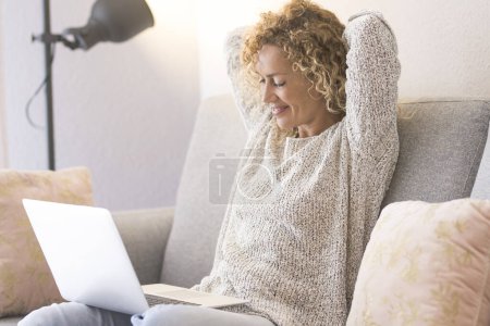 Téléchargez les photos : Femme utilisant un ordinateur portable assis sur le canapé dans la maison lumineuse. Les gens et la technologie en ligne. Femme moderne regardant l'ordinateur et détendre son dos et les épaules levant les bras sous la tête - en image libre de droit