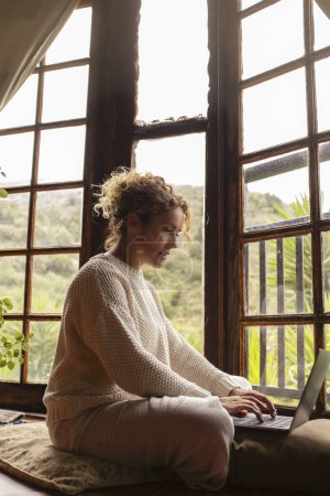 Photo for Woman sit down on the floor and use laptop in cozy wooden cabin with nature outdoors view. Concept of alternative healthy and technology lifestyle and small business. People using notebook indoor - Royalty Free Image