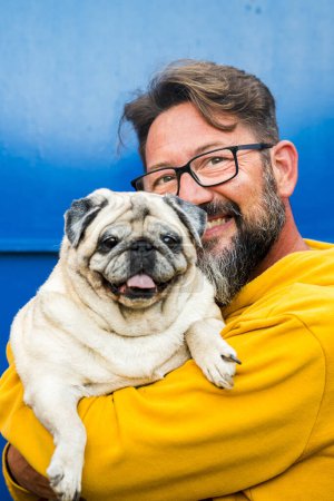Photo for Portrait of man and dog.  adult handsome with beard and eyewear hug and hold funny nice pug both looking on camera and blue background wall. Copy space and color. Love and friendship - Royalty Free Image