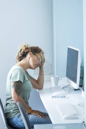 Photo for Stressed and tired adult  woman at the desktop in front of computer and online job activity day lifestyle touching her head for headache pain - office work - blue mood city colors - Royalty Free Image