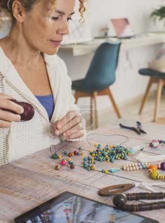 Foto de Busy woman at home start new small business doing jewelry with beads and using laptop to sell online or learn from tutorial. Indoor hobby leisure activity female people at the table alone - Imagen libre de derechos