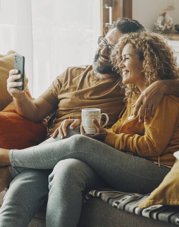 Foto de Couple of young adult man and woman enjoy time together sitting on the sofa in indoor leisure activity.. Real life people at home. Male and female relax on the couch reading a book and drinking a tea - Imagen libre de derechos