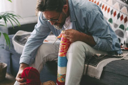 Foto de Man at home doing knit activity alone sitting on the sofa. Mature male people love knitting work for no stress hobby and relax time. Concept of person and leisure relaxed lifestyle indoor - Imagen libre de derechos