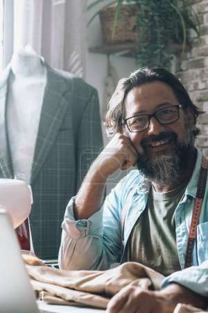 Photo for Portrait of a tailor sitting at the desk with laptop and sewing machine. Modern  tailor job and online store sell opportunity. Elegant blazer in background. People happy smiling and looking - Royalty Free Image