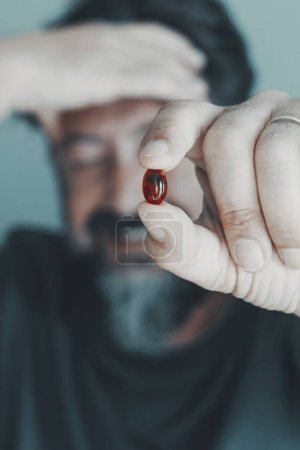 Téléchargez les photos : Gros plan de l'homme tenant la pilule rouge contre les migraines. Personne en arrière-plan tenant et touchant son front avec douleur. Concept de pharmacie et de traitement médical. Concentrez-vous sur les pilules - en image libre de droit