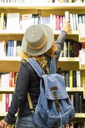 Téléchargez les photos : Back view of woman with hat and backpack choosing a book to buy inside a library or newspaper store. People prepare to travel. Leisure and shopping, female people activity. Traveler lifestyle - en image libre de droit