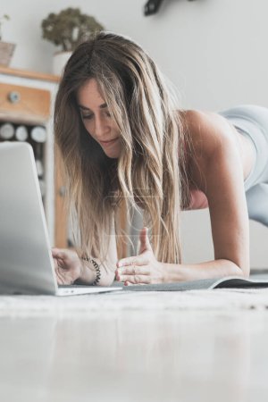 Photo for Young woman exercising at home doing push-ups and looking at her personal laptop computer to learn or teach workout - plank strength body shape workout online class alternative smart working people - Royalty Free Image