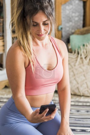 Photo for Shot of fitness woman sitting on yoga mat and using mobile phone. Fit young woman using cellphone while doing exercise at home. online class lesson sporty people with healthy lifestyle - Royalty Free Image