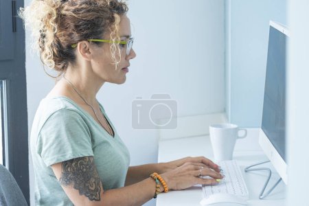 Photo for Cute modern style adult woman tattooed write and work on desktop computer in office or home room - concept of free people and online technology job activity - pretty adult lady use keyboard in white - Royalty Free Image