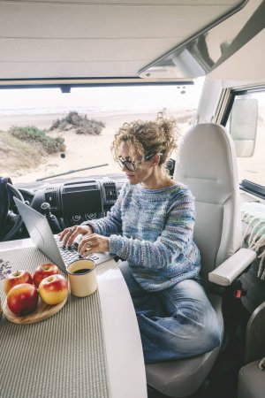 Photo for Living and working inside  camper while traveling and a digital nomad-free lifestyle. A woman sitting in an RV enjoys relaxing and connecting to her laptop. The beach in the background outside the window - Royalty Free Image