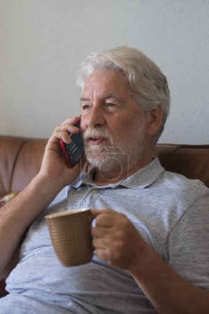Foto de Viejo hombre mayor en casa hablando con teléfono moderno y beber café o té sentado en el sofá y disfrutar de la conexión de la tecnología - un hombre maduro uso celular en casa relajarse y divertirse - Imagen libre de derechos