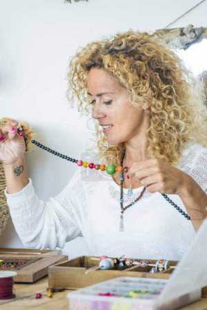 Photo for Happy woman at homework with beads and cords producing cheaper jewelry-modern female alternative job at home at the table - colorful trendy fashion young female - Royalty Free Image