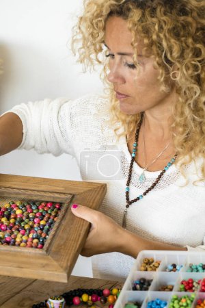 Photo for Adult woman doing hand made creations jewelry at home with colorful beads and cords - female people working at home for online store business shopping - handcraft diy hobby female and jewels on a wooden table - Royalty Free Image
