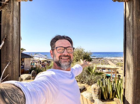 Téléchargez les photos : Adulte homme heureux prenant des photos selfie avec téléphone appareil photo dans une activité de loisirs de plein air avec bar de plage et océan en arrière-plan. Tourisme et voyage concept de destination mode de vie personnes. Joyeux mâle - en image libre de droit
