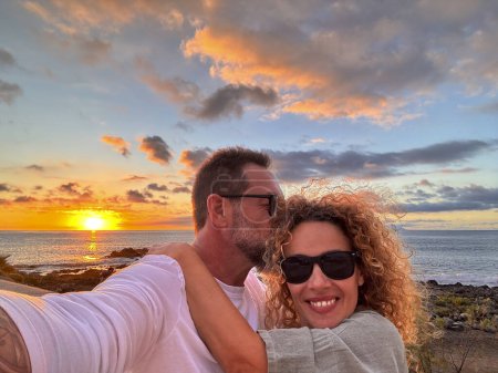 Téléchargez les photos : Romantic couple of man and woman in love taking selfie picture together hugging and smiling at the phone camera and enjoying beautiful sunset on the ocean at the beach. Travel people summer vacation - en image libre de droit