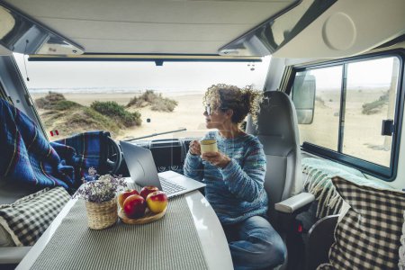 Foto de Vivir y trabajar dentro de su caravana mientras viaja y un estilo de vida digital libre de nómadas. Una mujer sentada en una caravana disfruta de relajarse y conectarse a su computadora portátil. La playa en el fondo fuera de la ventana - Imagen libre de derechos