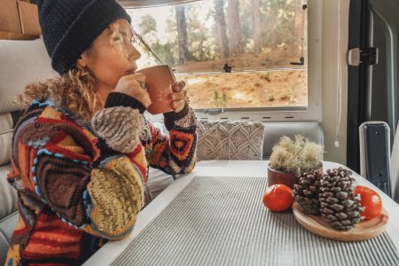 Photo for Woman with hat and patchwork sweater sitting in camper drinking hot tea looking out the window. Relaxation and leisure concept - Royalty Free Image