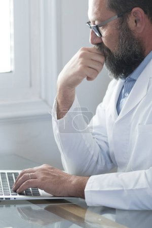 Photo for Adult mature Caucasian male doctor in white medical uniform sit at desk in office consult client on laptop online, man GP or physician fill patient medical card use Web application form on computer - Royalty Free Image
