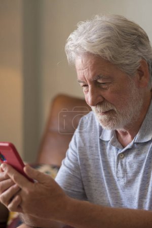 Photo for Mature handsome man text and write message on the smartphone - elderly people use cellular to chat sit down on the sofa at home - one retired people indoor lifestyle alone with connected device - Royalty Free Image