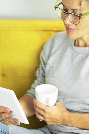 Photo for Close up portrait of mature pretty woman reading a book on tablet pc with glasses sitting on the yellow couch at home in break leisure time activity alone - adult people enjoy technology and learning - Royalty Free Image