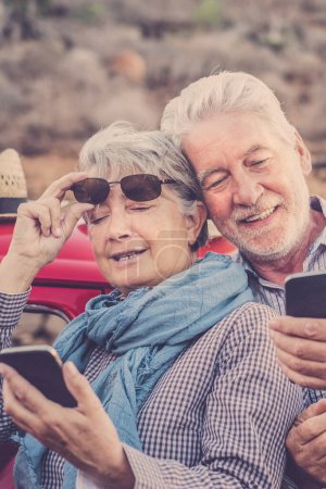 Téléchargez les photos : Joyeux vieux couple de personnes âgées utilisent les téléphones ensemble en plein air avec voiture rouge en arrière-plan concept de voyage les gens matures et la technologie moderne chez les personnes âgées retraitées mode de vie heureux - en image libre de droit